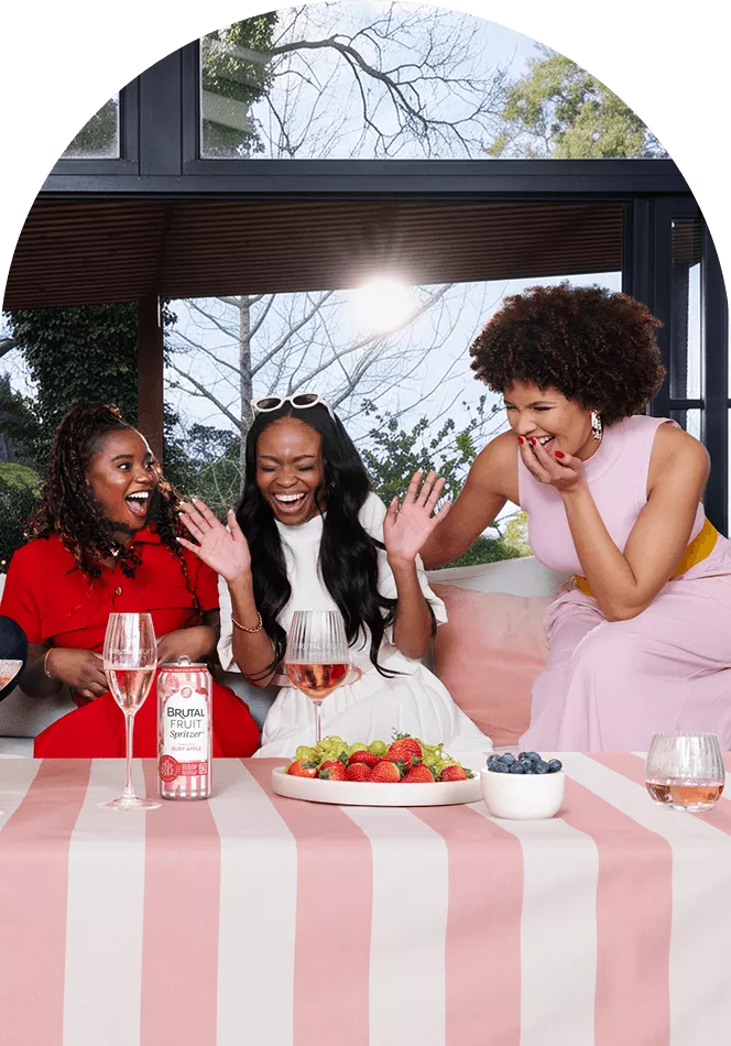 Three friends laughing at the table, with glasses, fresh fruit, and Brutal Fruit Spritzer.